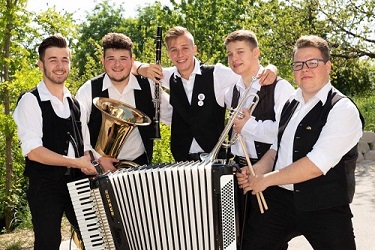 Öt fiatal zenész mellényben, harmonikával és rézfúvós hangszerekkel a szabadban.