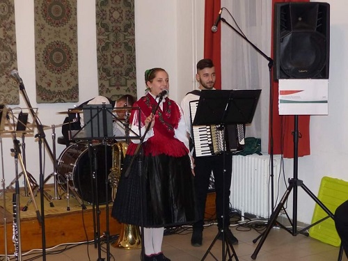 Népviseletbe öltözött énekesnő és harmonikás zenél a Német Ház termében.