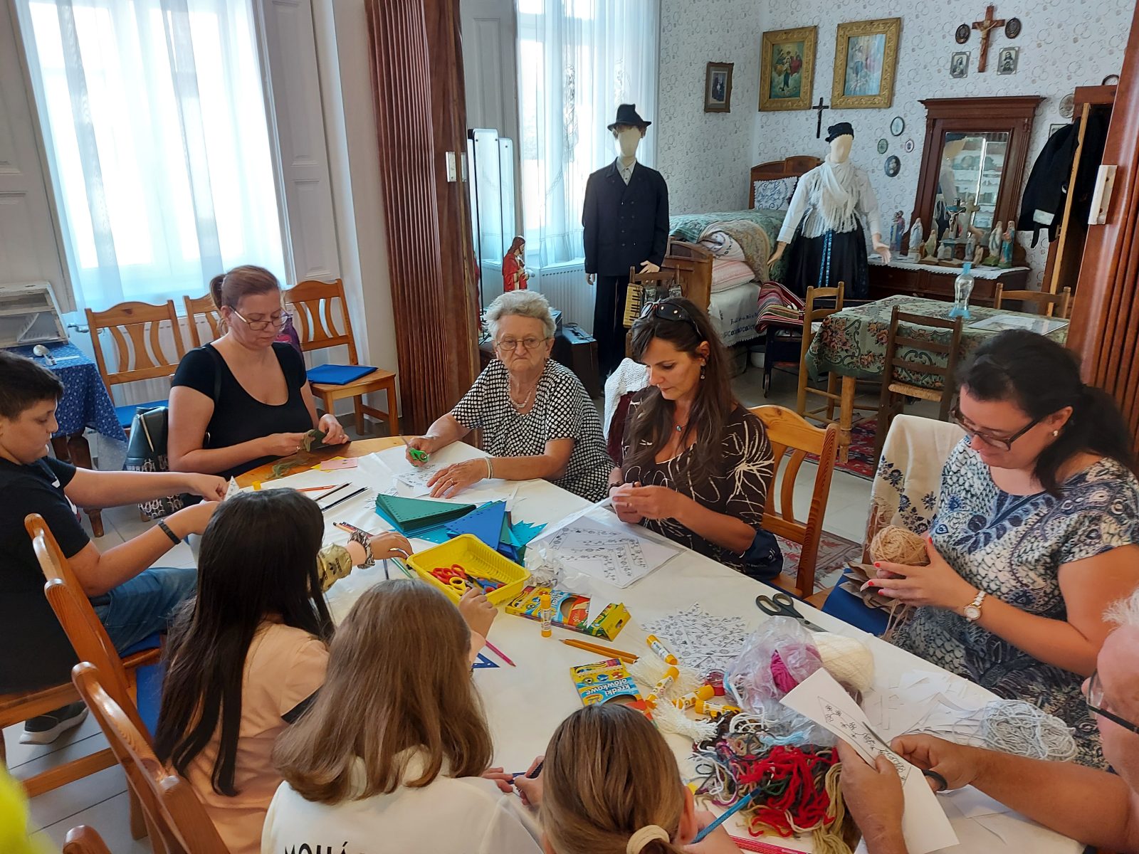 Felnőttek és gyerekek kézimunkáznak egy hosszú asztal körül, a háttérben népviseletbe öltöztetett próbababák.