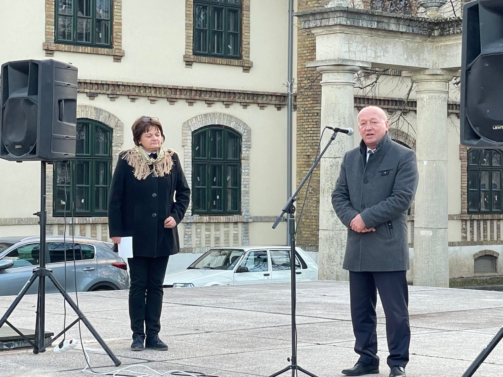 Nő és férfi beszédet mond mikrofon előtt egy régi épület homlokzata előtt.