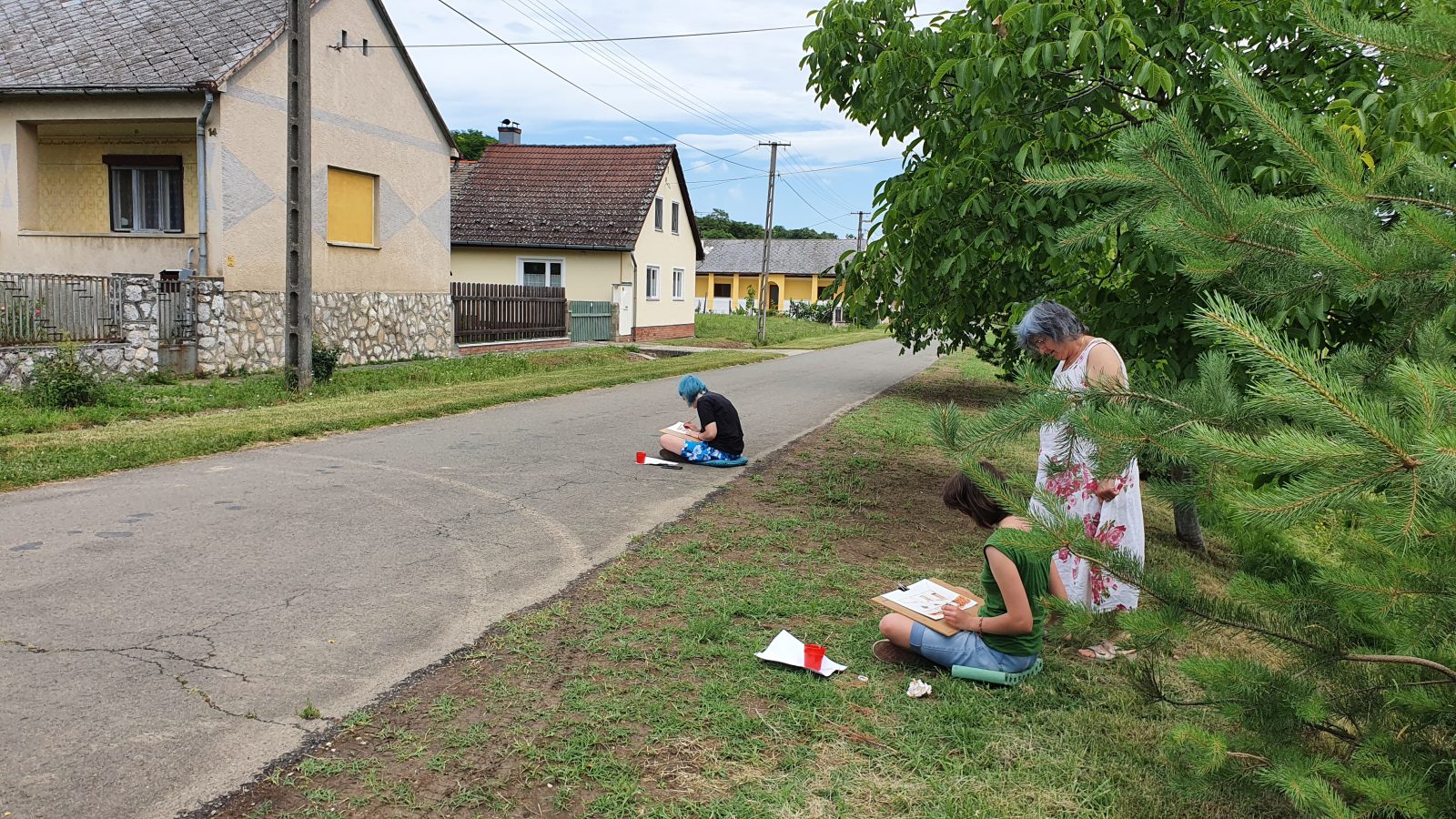 Alkotótábor résztvevői rajzolnak az út menti füvön egy falusi utcában.