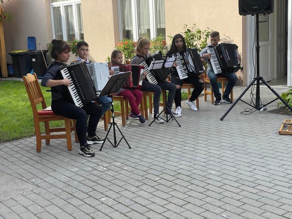 Fiatalok harmonikáznak a szabadban, székeken ülve.