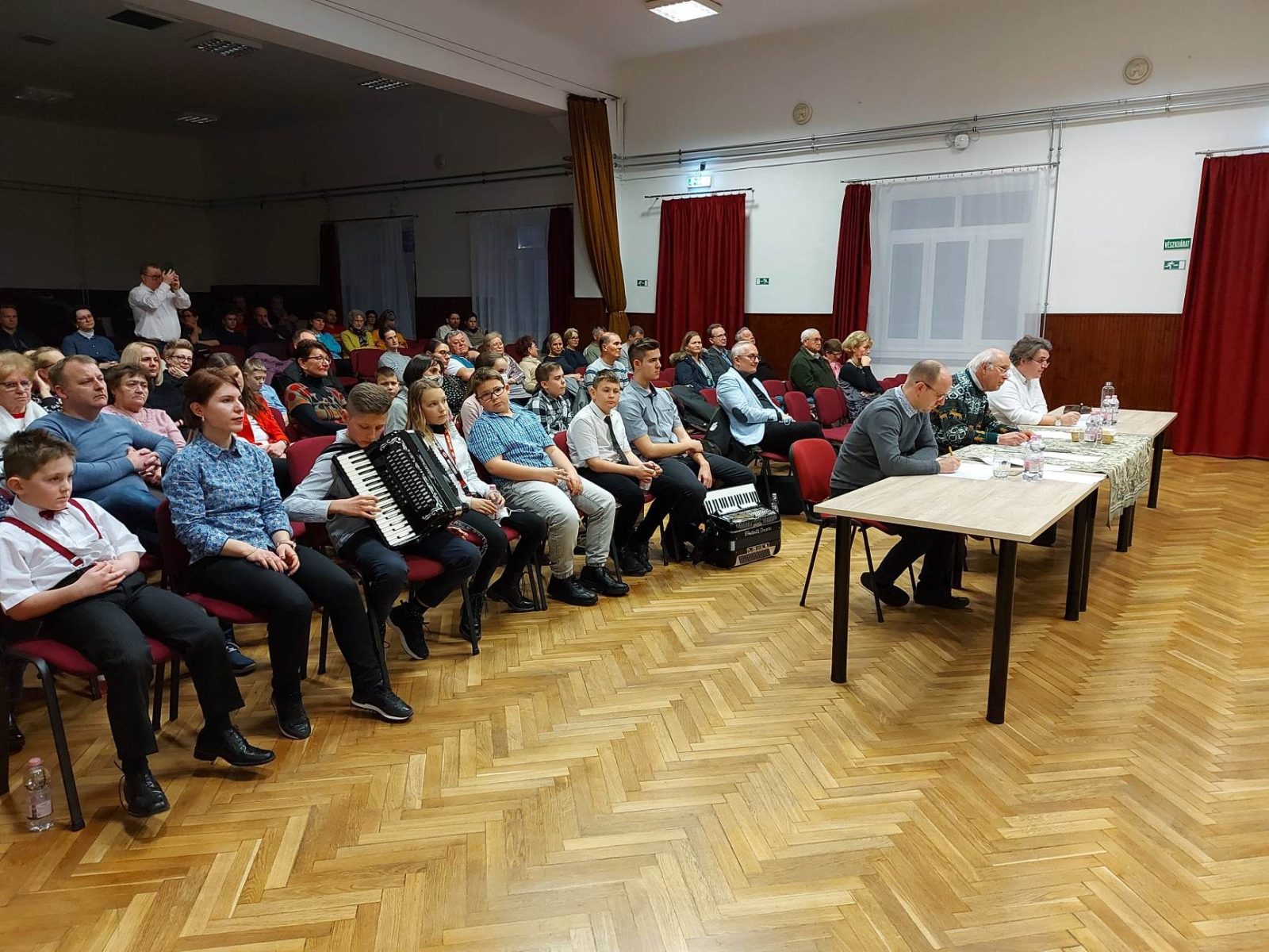 Megtelt terem harmonikás versenyzőkkel és a zsűri asztalával.