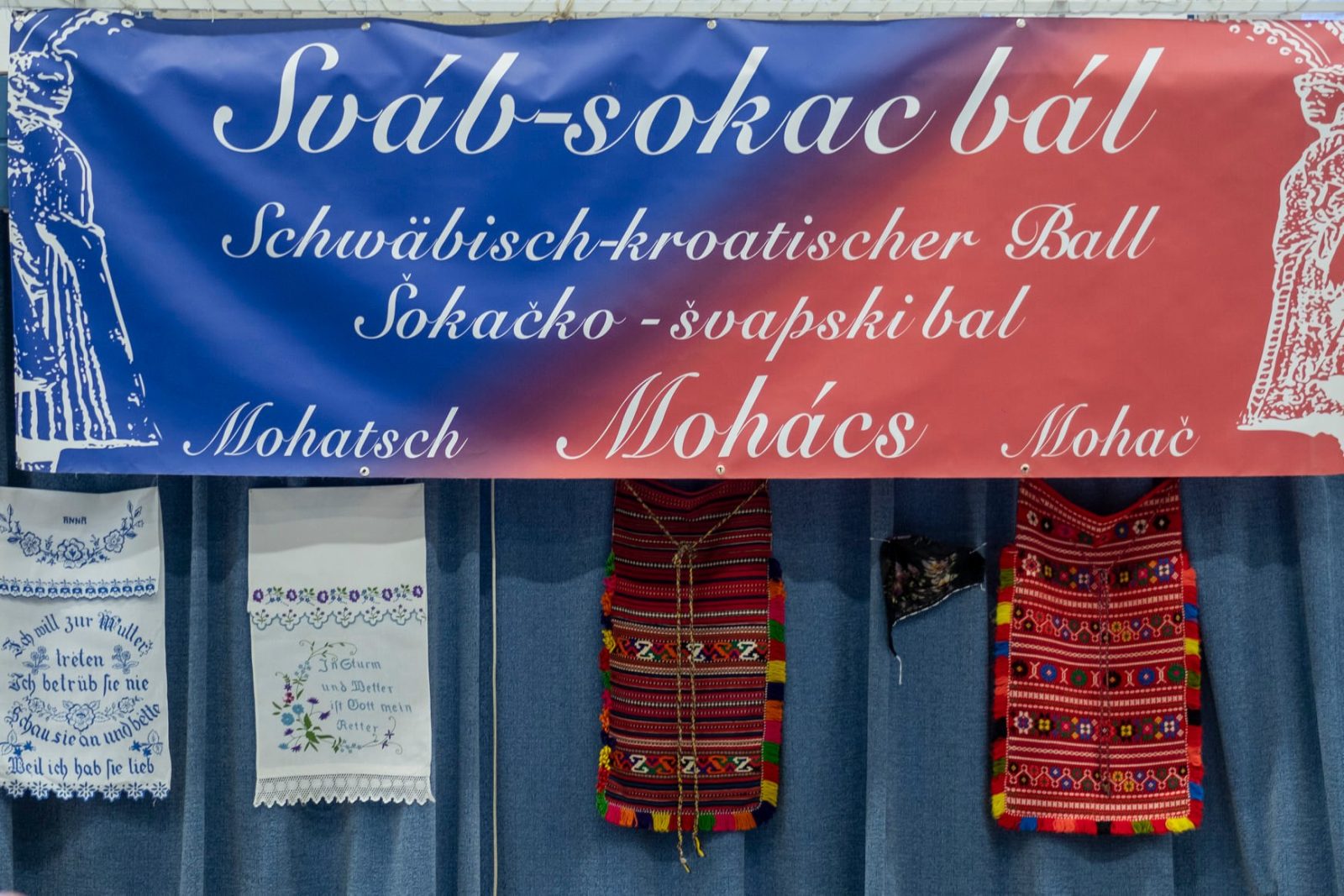 „Sváb-sokac bál” feliratú molinó, alatta hímzett terítők és népviseleti ruhadarabok.
