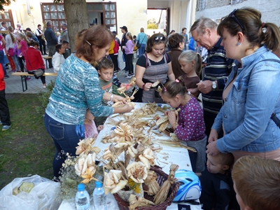 Gyerekek és felnőttek kukoricacsuhéból készítenek figurákat egy hosszú asztalnál.