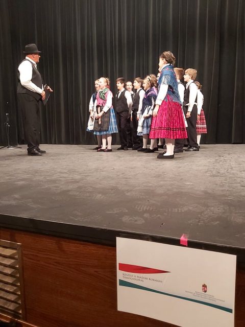 Gyerekek népviseletben a színpadon, mellettük kalapos férfi.