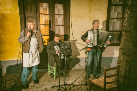Énekes és két harmonikás lép fel egy régi ház tornáca előtt.