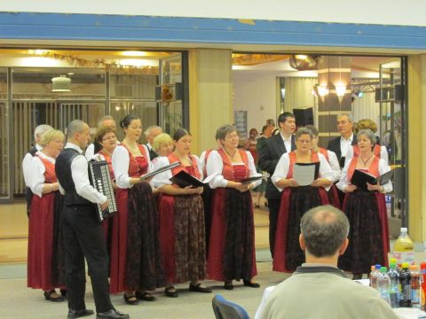Népviseletbe öltözött énekkar harmonikás kísérővel egy teremben.