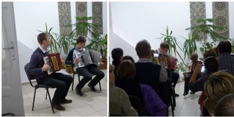 Harmonikázó férfiak egy növényekkel teli teremben, előttük hallgatóság.