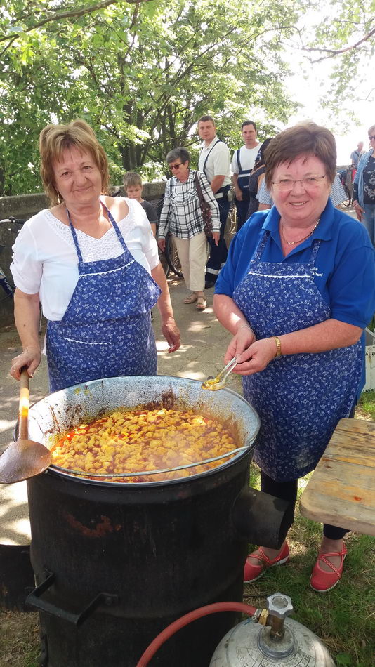 Két asszony kötényben nagy bográcsban főz a szabadban.