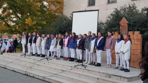 Nagy csoport énekel a szabadban egy téglafal előtt, mikrofonokkal.