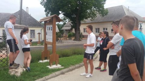 Fiatalok egy tájház udvarán, mellettük fából ácsolt építmény.