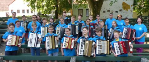 Kék pólós fiatalok nagy csoportja harmonikákkal a szabadban.