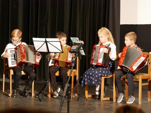 Négy gyerek harmonikázik a színpadon, kottaállványok mögött.