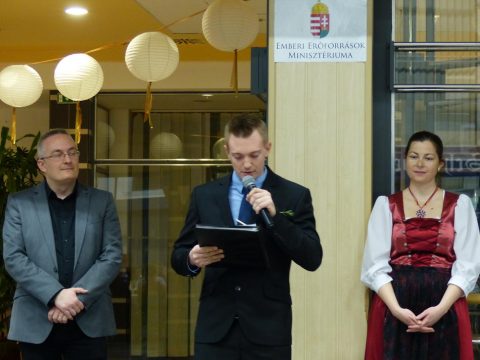 Öltönyös férfi mikrofonnal beszél, mellette egy férfi és egy népviseletes nő.