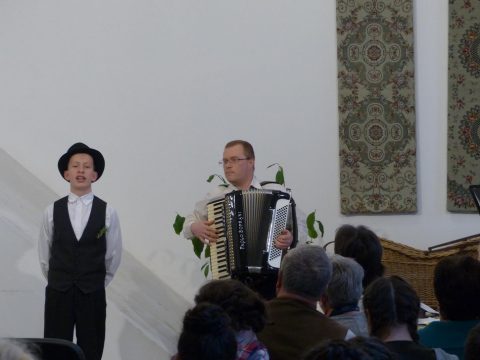 Kalapos énekes és harmonikás lép fel a színpadon a közönség előtt.