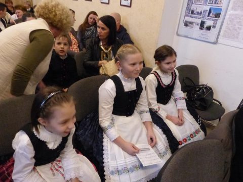 Két kislány sváb népviseletben ül a nézőtéren, kezükben papírral.