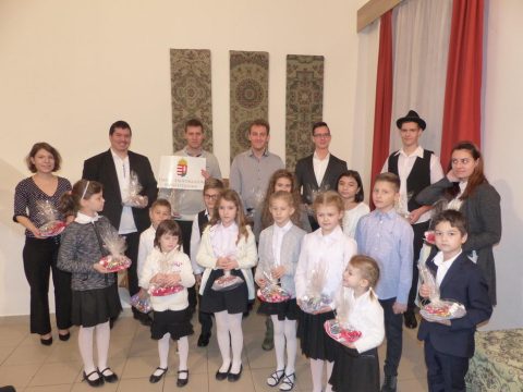 Fehér inges gyerekek és felnőttek csoportja tányérokkal a színpadon, a háttérben szőttesek.