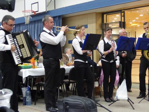 Zenészek harmonikával, trombitával és klarinéttal játszanak egy asztal mellett.